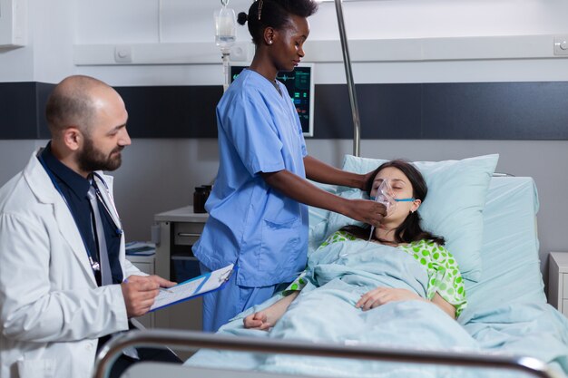 Afro american asisstant putting oxigen mask to sick woman while lying in bed having respiratory dise...