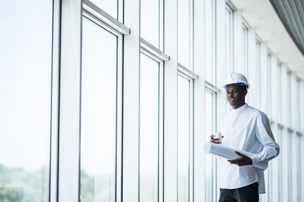 Afro american construction engineer in front of building holding blueprints against windows