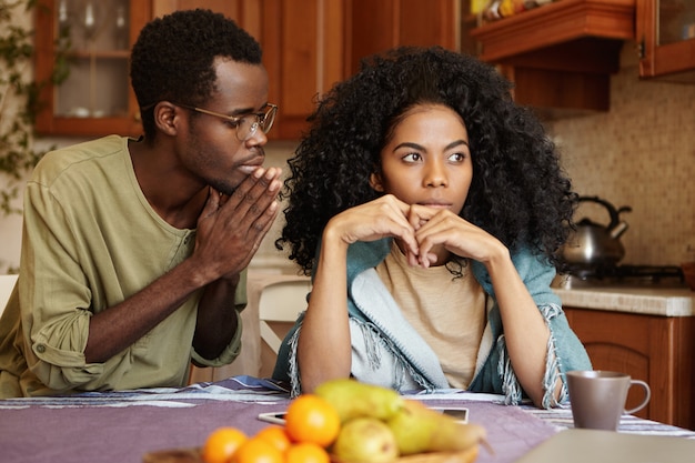 Afro-american couple going through hard times in their relationships. guilty unfaithful young man keeping hands pressed begging his angry wife to forgive him for infidelity, trying to sweet talk her