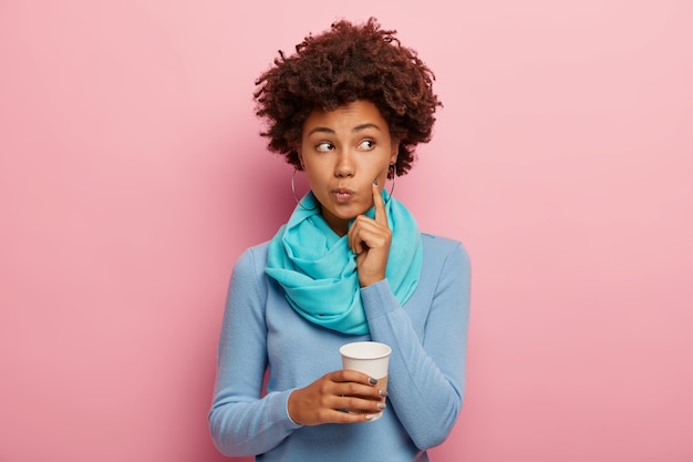 Afro american curly woman keeps index finger on cheek, looks thoughtfully aside, contemplates about something with hot drink, holds paper cup, wears blue jumper, has coffee break isolated on pink wall
