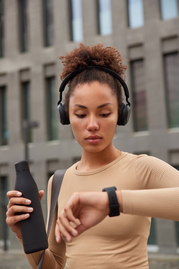 Free Photo afro american female athlete drinks fresh water afterworkout checks heartbeat on smartwatch listens music in headphones poses in city