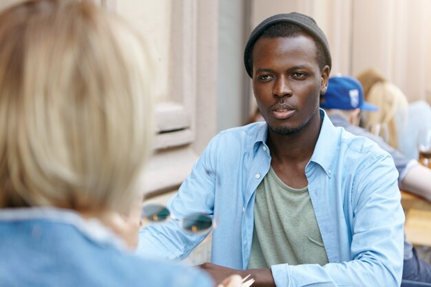Afro American guy with dark skin dressed in shirt and black hat sitting in front of his female friend, having conversation with each other, discussing news. Business partners meeting at cafe