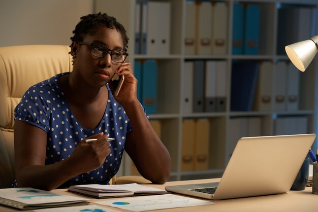 Afro-American lady sitting at desk in office at night and talking on mobile phone