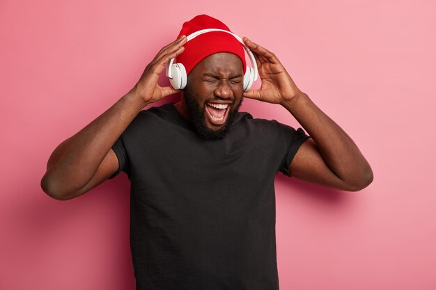 Афро-американец наслаждается музыкальным плеером или аудиозаписью, держит в руках наушники, развлекается, слушая популярные песни, изолированные на розовой стене.