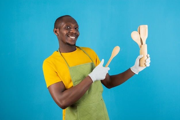 Afro american man in green apron holding kitchen wooden tools on blue wall