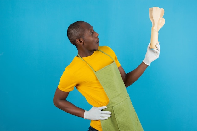 Афро-американский мужчина в зеленом фартуке держит кухонные деревянные инструменты на синей стене
