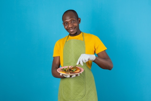 青い壁に食べ物のプレートを保持している緑のエプロンのアフロアメリカ人