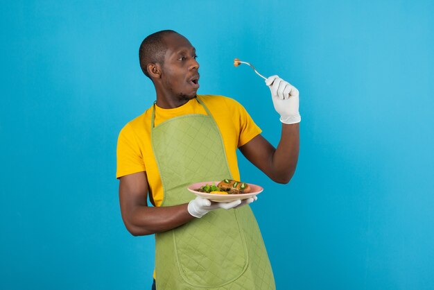 Афро-американский мужчина в зеленом фартуке держит тарелку с едой на синей стене