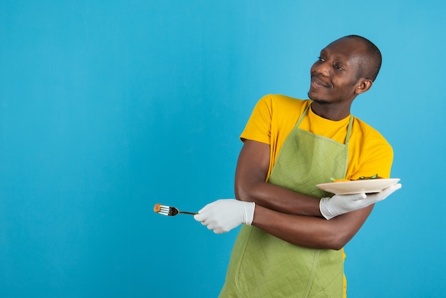 Афро-американский мужчина в зеленом фартуке держит тарелку с едой на синей стене