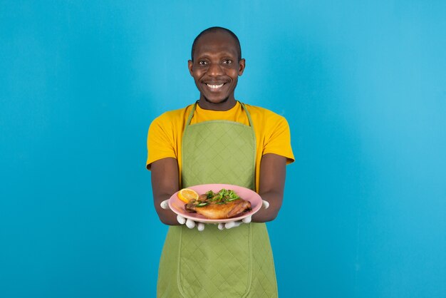 Afro american man in green apron offering plate of food on blue wall