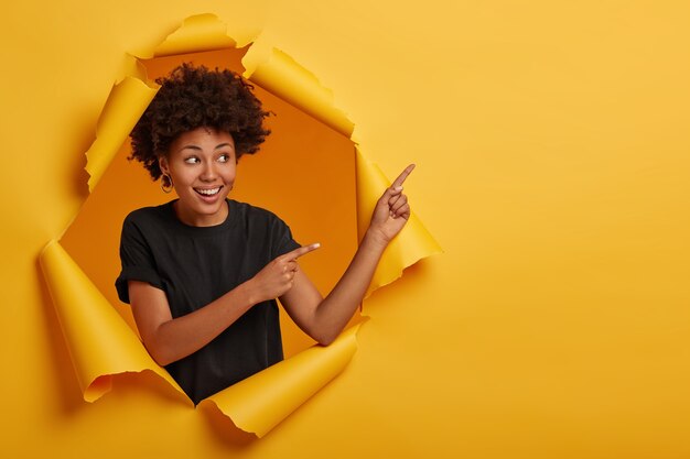 Afro American woman checks out incredible offer, points right with index fingers, suggests buying something