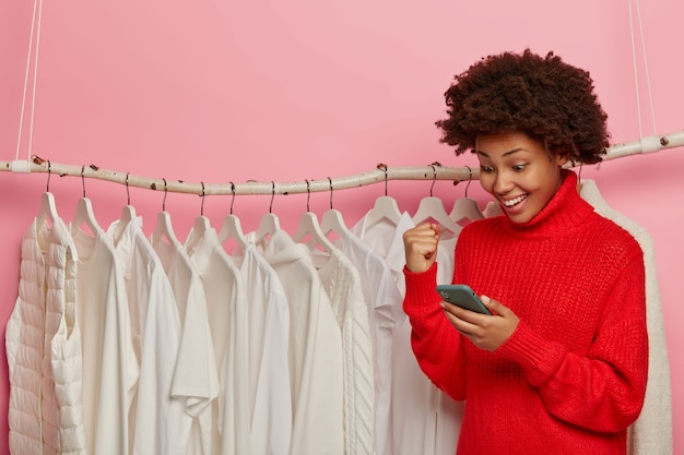 Free photo afro american woman cheers something with clenched fist, looks positively at smartphone, poses against clothes rails with white outfits on hangers, makes shopping in boutique