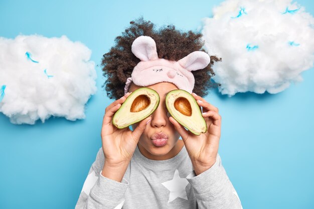 Афро-американка закрывает глаза половинками авокадо, держит губы округлыми, собирается сделать питательную маску для лица, носит пижаму и маску для сна, изолированные на синем