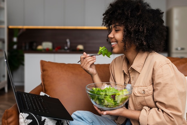 Afro-american woman eating salad and looking on laptop