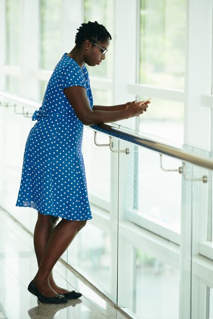 Afro-American woman in polka-dot dress leaning on handrail on balcony and using smartphone