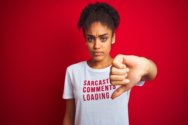 우스꽝스러운 티셔츠를 입은 아프로계 미국인 여성은 격리된 빨간색 배경 위에 반어적인 댓글을 달고 엄지손가락을 아래로 거부하는 개념으로 싫어하는 것을 보여주는 화난 얼굴 부정적인 기호를 가지고 있습니다.