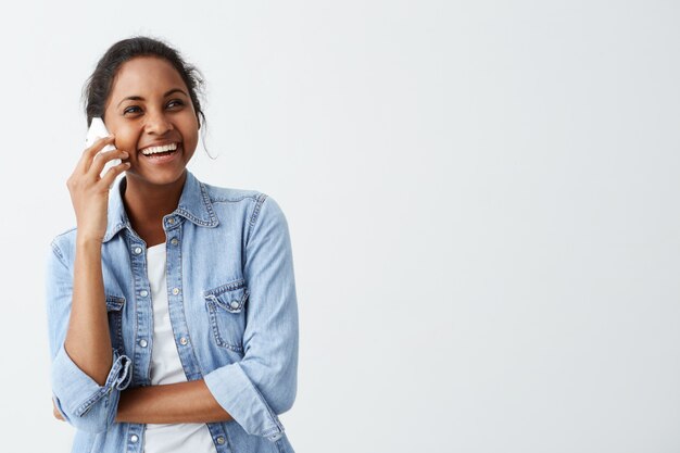 アフリカ系アメリカ人の若い嬉しい女性が白いtシャツに青いシャツを着て、スマートフォンで会話し、笑いながら、友達と良いニュースを共有しています。人と前向きな感情。