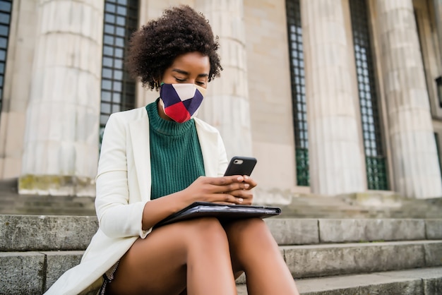 Afro business woman wearing protective mask and using her mobile phone while sitting on stairs outdoors at the street. Business and urban concept.