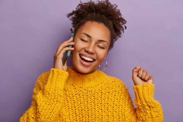 アフロの女性は電話で会話し、面白い面白い話をし、くいしばられた握りこぶしを上げ、紫色の背景の上に広く孤立した笑顔を持っています