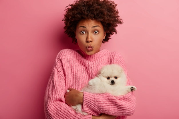 Afro woman keeps lips folded, carries little Pomeranian spitz to grooming salon, cares about animal, wears pink jumper