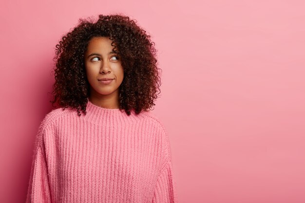 Afro woman thinks deeply about something, thinks how to act in difficult situation, wears pink knitted sweater stands indoor