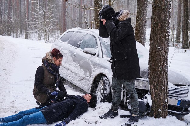 После автомобильной аварии в зимнем лесу раненый мужчина лежит на снегу, женщина пытается ему помочь, еще один мужчина в отчаянии.