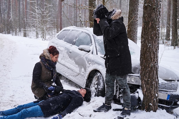 冬の森で車が押しつぶされた後、負傷した男性が雪の上に横たわっていた後、女性は彼を助けようとしています、別の男性は絶望しています。