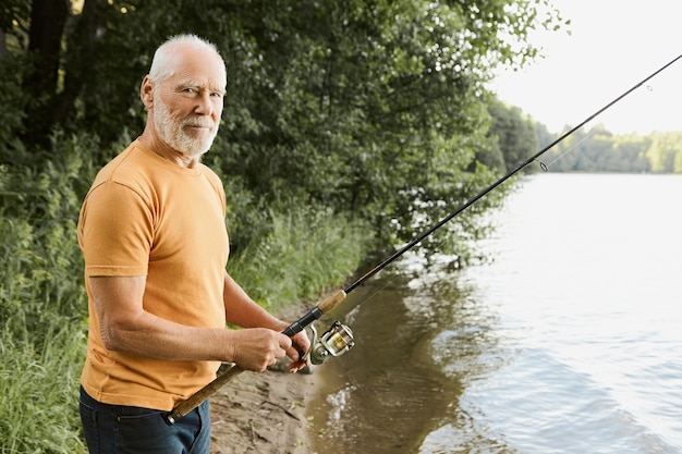 年齢、活動、レジャーの概念。釣り竿を水に投げ込み、魚が引っ掛かるのを待って川岸で釣りをしている間、リラックスして幸せを感じている引退したシニアひげを生やした男性の側面図