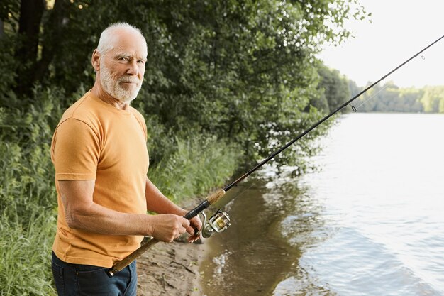 Age, activity and leisure concept. Side view of retired senior bearded male feeling relaxed and happy while fishing on river bank with fisher rod cast in water, waiting for fish to be hooked