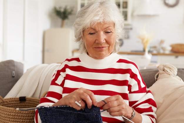 Foto gratuita età, tempo libero, hobby e concetto di pensionamento. pensionato femminile caucasico elegante attraente in maglione rosso bianco a strisce che si siede sul divano in interni accoglienti con aghi e filati, sciarpa o cappello per maglieria