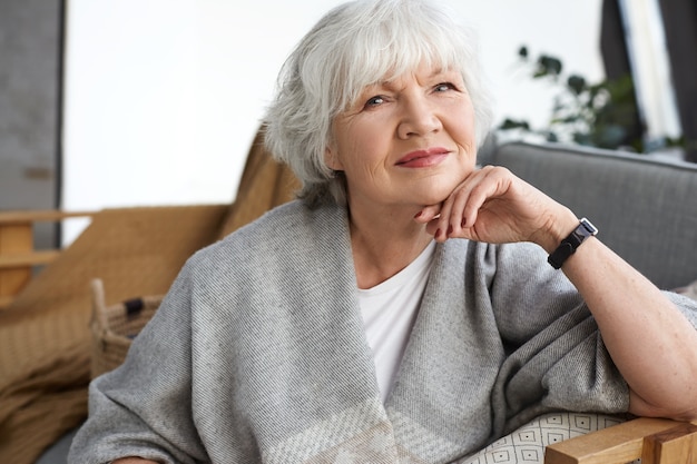 Age, retirement and relaxation concept. Happy cheerful mature retired woman looking with beaming smile, enjoying nice winter day, sitting on couch, wrapped in wide scarf, dreaming