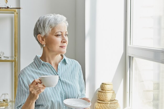 Age, style and maturity concept. Beautiful stylish middle aged woman with gray pixie hair enjoying herbal tea, holding white mug and saucer, looking through window, having thoughtful facial expression