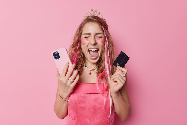 Age of technology Emotional young woman shouts loudly keeps mouth widely opened poses with smartphone and credit card uses modern gadget to pay for goods in online store wears crown and dress