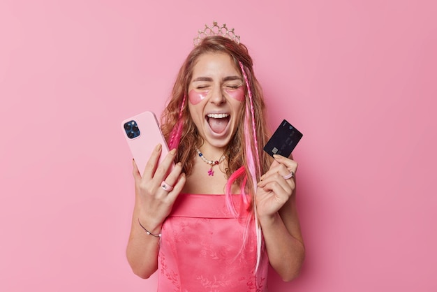 Эпоха технологий Эмоциональная молодая женщина громко кричит, широко раскрывает рот, позирует со смартфоном и кредитной картой, использует современный гаджет для оплаты товаров в интернет-магазине, носит корону и платье