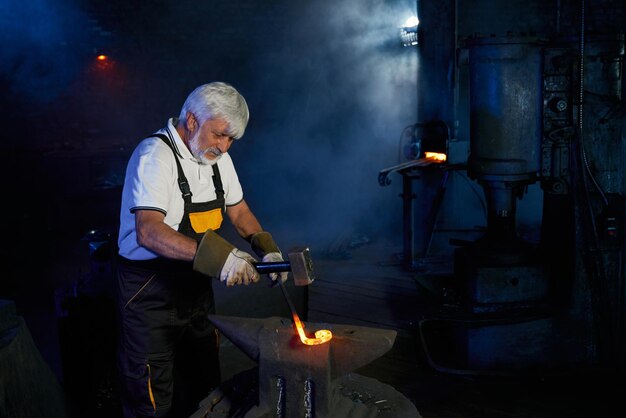 Aged caucasian blacksmith wearing safety apron and gloves forging steel on anvil with heavy hammer Manual work at forge Manufacturing concept