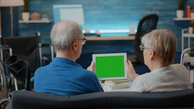 Aged couple with chronic disability using green screen on digital tablet. Senior people looking at isolated chroma key with blank mock up template and copy space background on device.