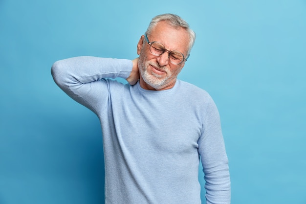 Aged exhausted bearded European man touches neck suffers from pain in neck tilts head grimaces from painful feelings needs massage dressed in long sleeved jumper isolated over blue wall