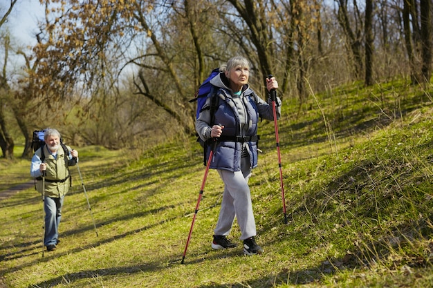 Пожилая семейная пара мужчина и женщина в туристическом наряде гуляют по зеленой лужайке в солнечный день