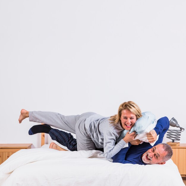 Aged happy woman lying on man with pillows on bed in bedroom