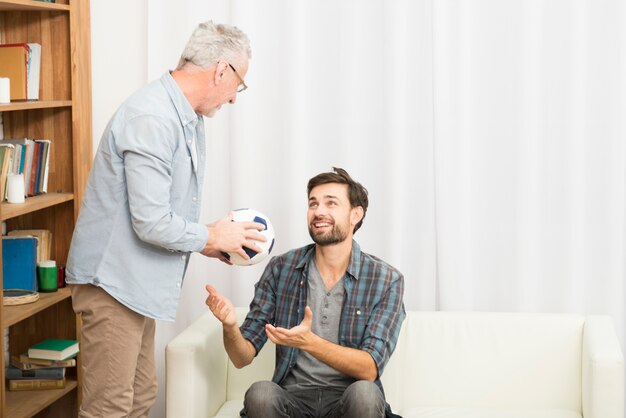 Aged man giving ball to young guy on sofa