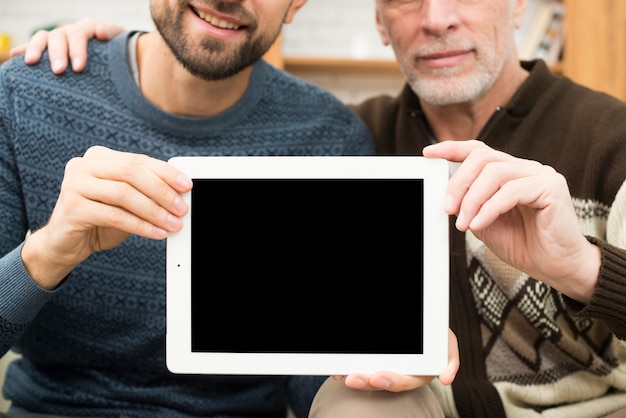 Aged man hugging young guy and showing tablet