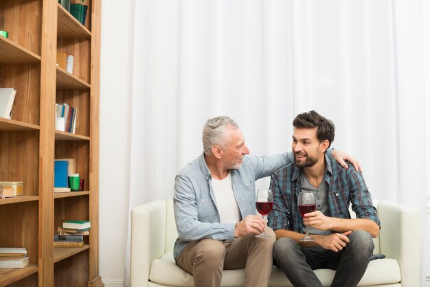 Пожилой мужчина обнимает молодого улыбающегося парня с бокалами вина на диване в комнате