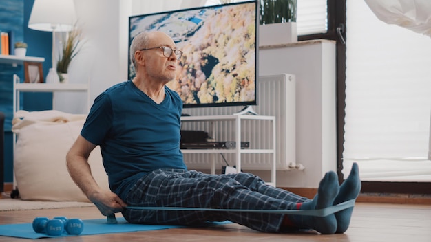 Пожилой мужчина использует эластичную ленту, чтобы растянуть руки и ноги на коврике для йоги. Пожилой человек тянет гибкий эластичный пояс, растягивает мышцы и делает фитнес-упражнения. Обучение взрослых старшего возраста