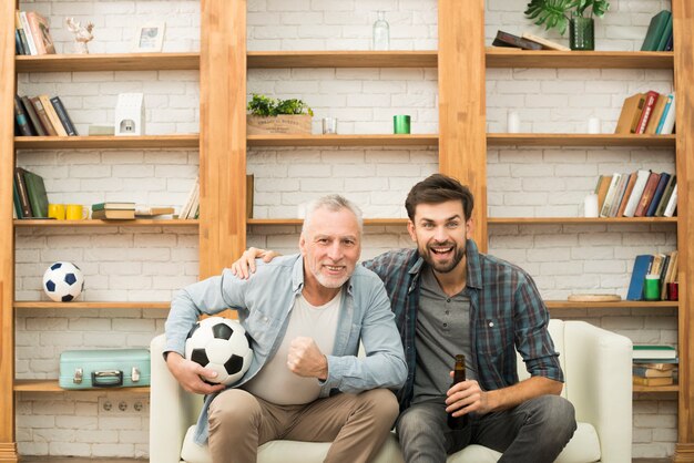 Aged man with ball and young guy with bottle watching TV on sofa