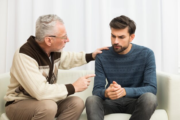 Aged man with hand on shoulder pointing at young guy on settee