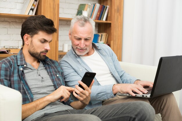 Aged man with laptop and young guy using smartphone on sofa