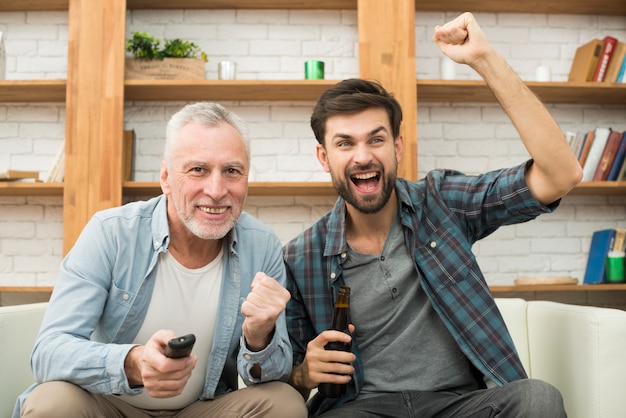Aged man with remote control and young crying guy with bottle watching TV on sofa