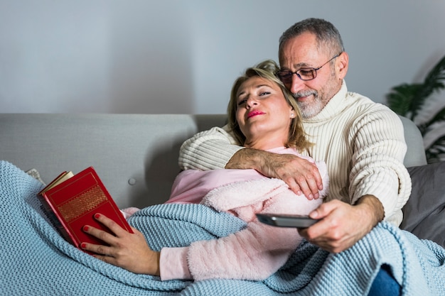 Aged man with TV remote embracing woman with book on sofa