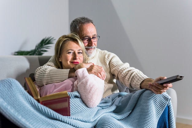 Aged man with TV remote watching TV and smiling woman with book on sofa