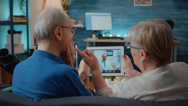 老人と女性がオンラインビデオ通話で歯科医と話し、歯痛について尋ねています。ビデオ電話会議を使用してデジタルタブレットで歯科矯正医とチャットする高齢者。リモート遠隔医療
